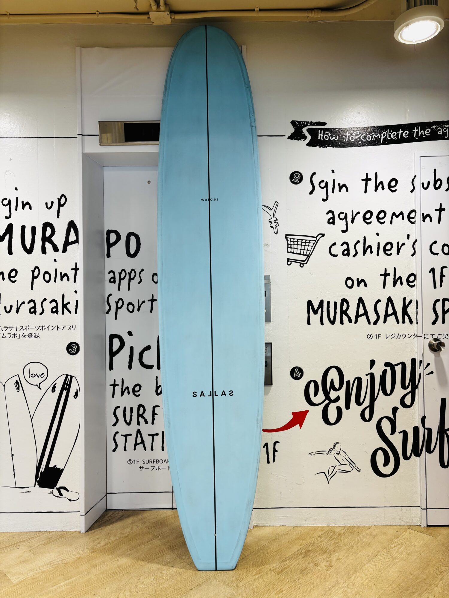 IMG_0480 - SHOP NEWS | ムラサキスポーツオンラインストア 通販 Blue surfboard leans upright against a white wall covered with black handwritten text and drawings, in a room with a wooden floor.