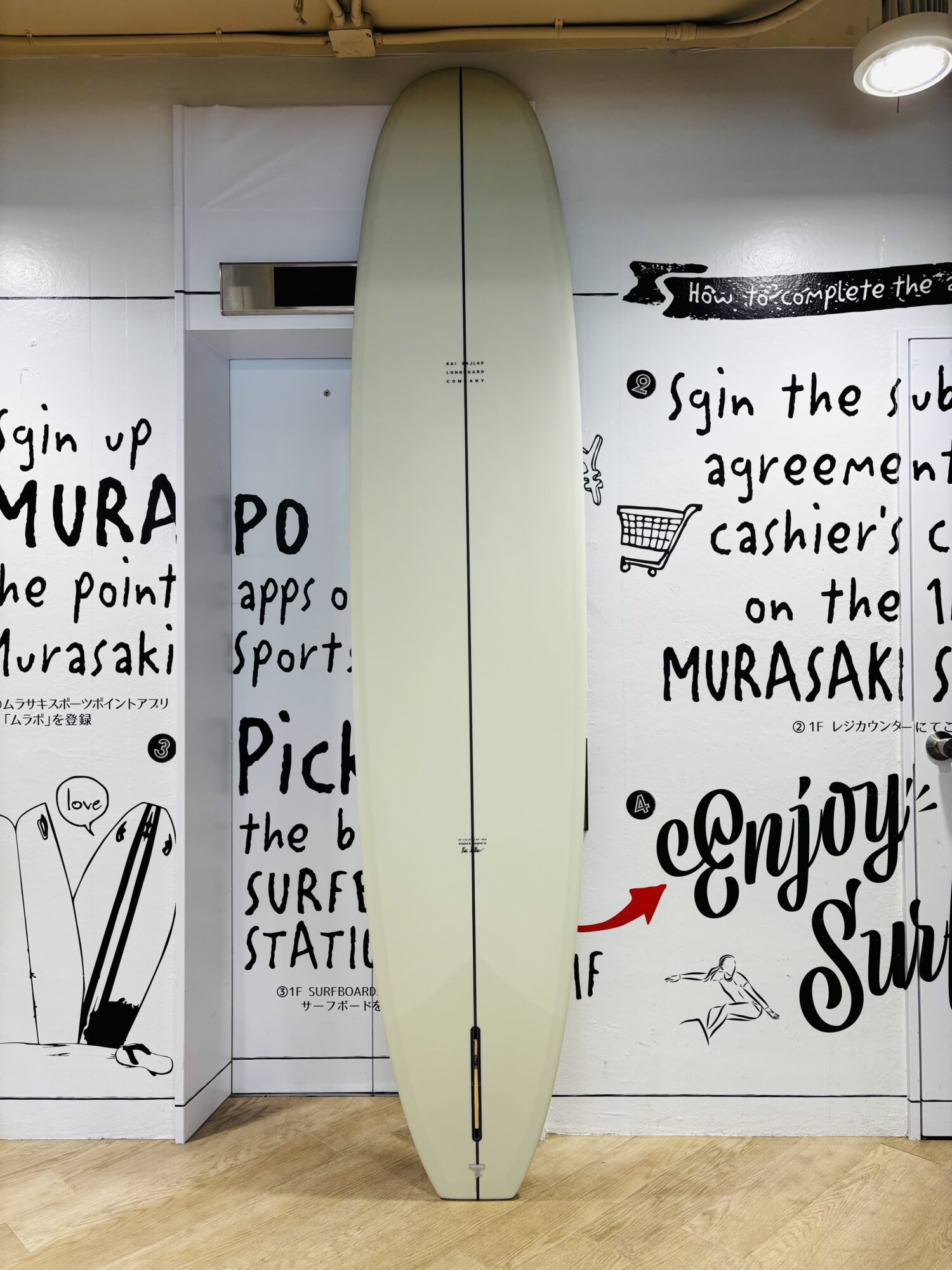 IMG_8974 - SHOP NEWS | ムラサキスポーツオンラインストア 通販 Cream-colored surfboard standing upright against a white wall with bold black graffiti text indoors, with warm overhead lighting and a wooden floor.