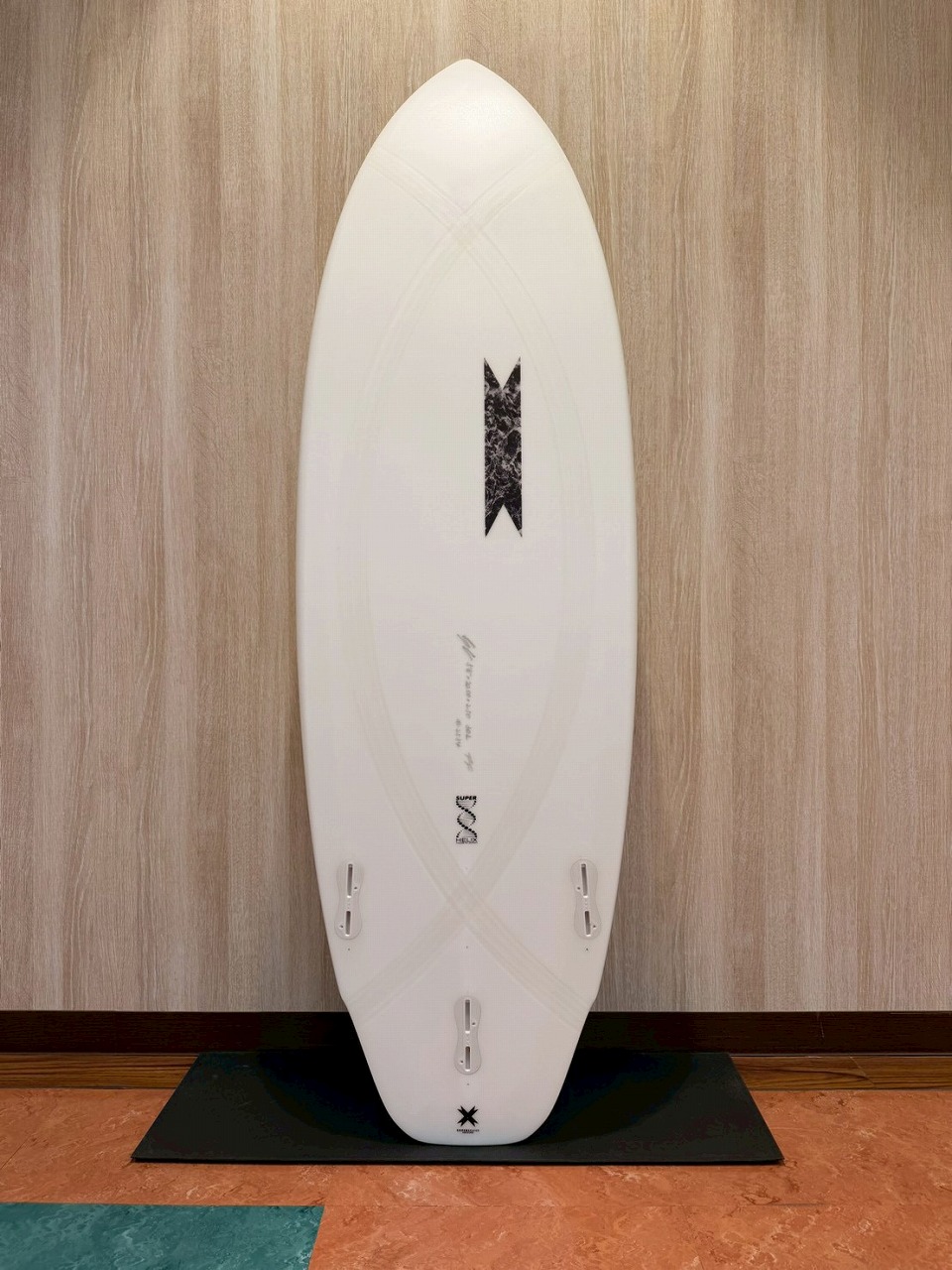 White surfboard standing upright on a black mat against a wood-paneled wall; three fin boxes visible near the bottom.