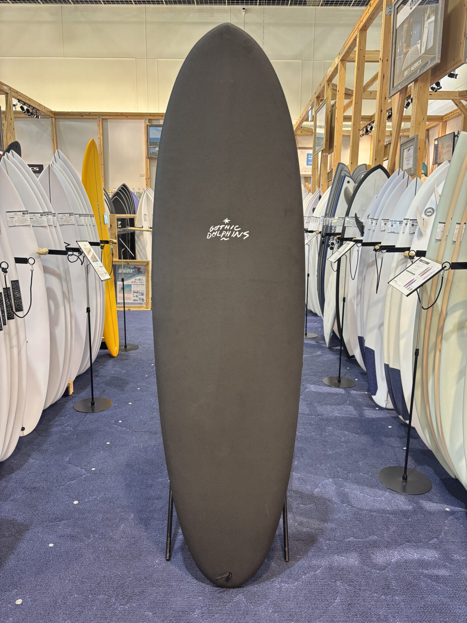 IMG_4172 - SHOP NEWS | ムラサキスポーツオンラインストア 通販 Tall black surfboard with Gothic Dolphins logo displayed upright in a showroom, surrounded by rows of white and colored boards on racks.