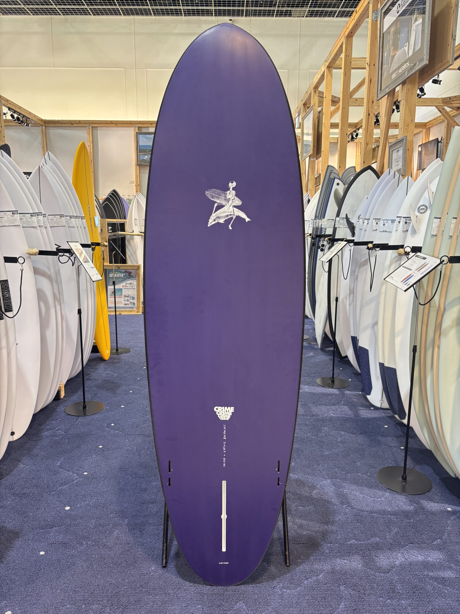 IMG_4173 - SHOP NEWS | ムラサキスポーツオンラインストア 通販 Tall purple surfboard on a stand in a showroom, surrounded by white and colored surfboards on racks.
