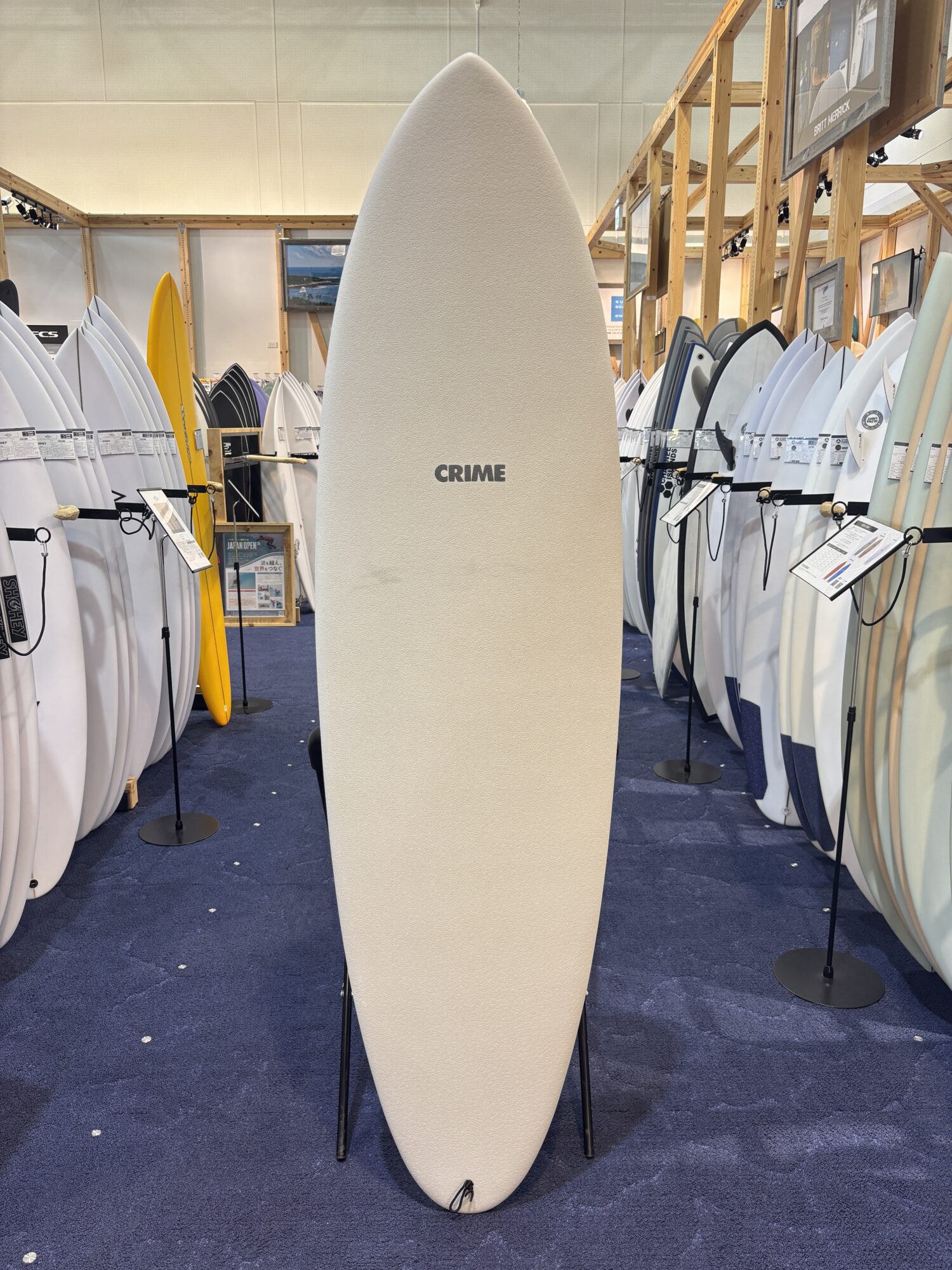 IMG_4174 - SHOP NEWS | ムラサキスポーツオンラインストア 通販 Large beige surfboard stands upright in a showroom, surrounded by rows of other surfboards on racks; the center board has 'CRIME' written on it.