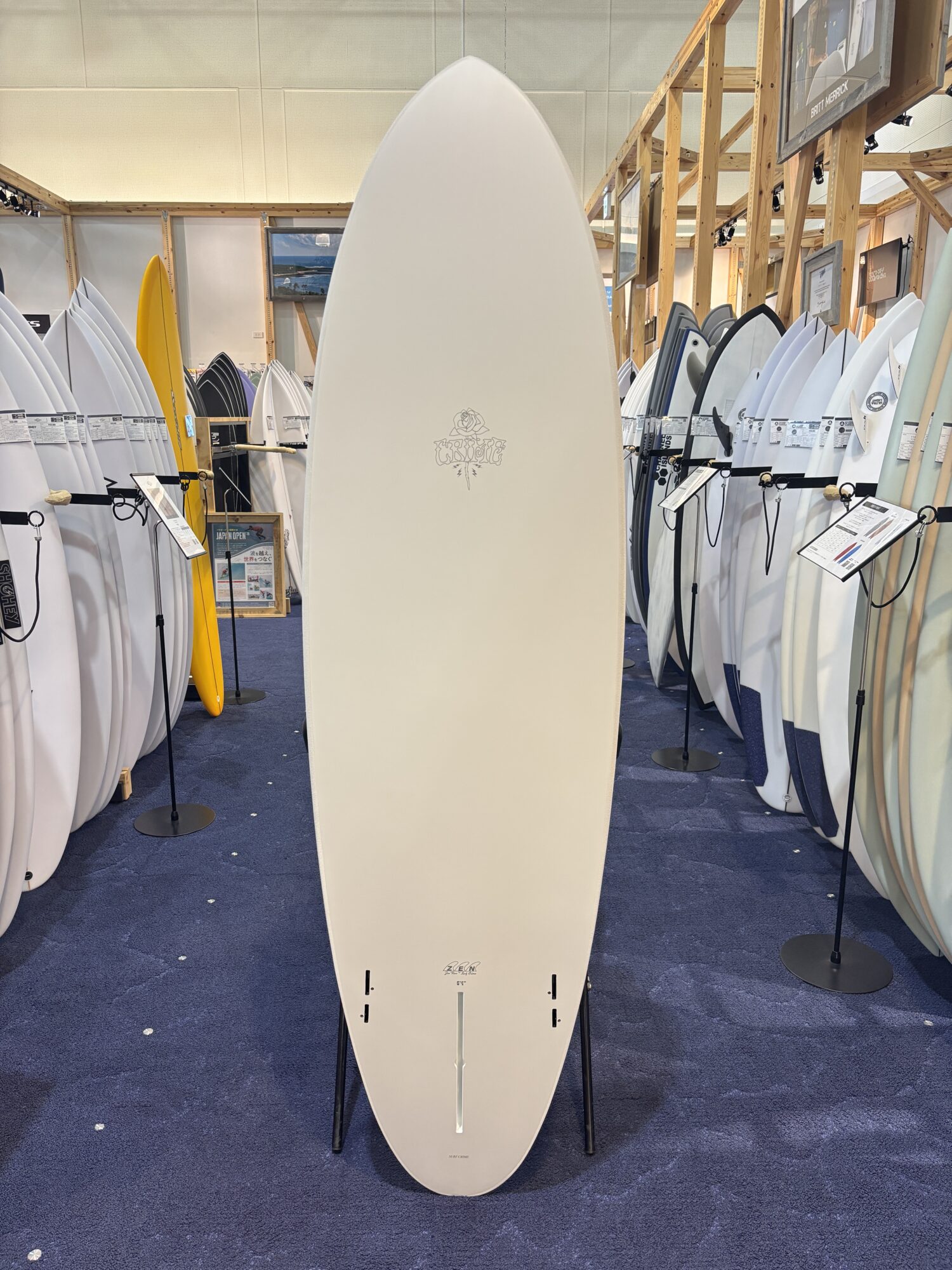 IMG_4175 - SHOP NEWS | ムラサキスポーツオンラインストア 通販 Tall white surfboard displayed upright on a stand in a showroom, surrounded by many other surfboards of various colors and sizes on racks.