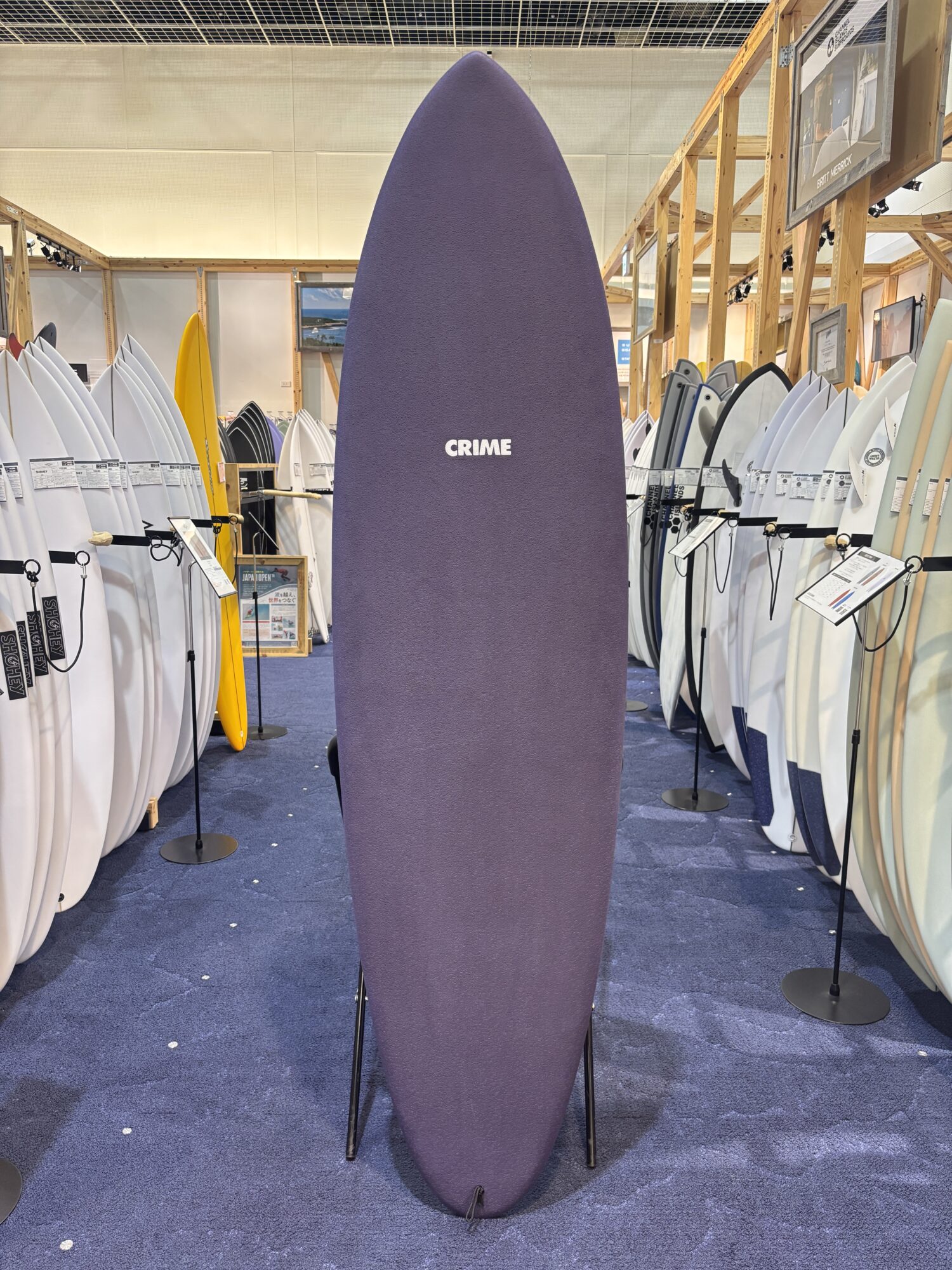 IMG_4177 - SHOP NEWS | ムラサキスポーツオンラインストア 通販 Tall purple surfboard displayed upright in a showroom, flanked by white and colored boards on racks.