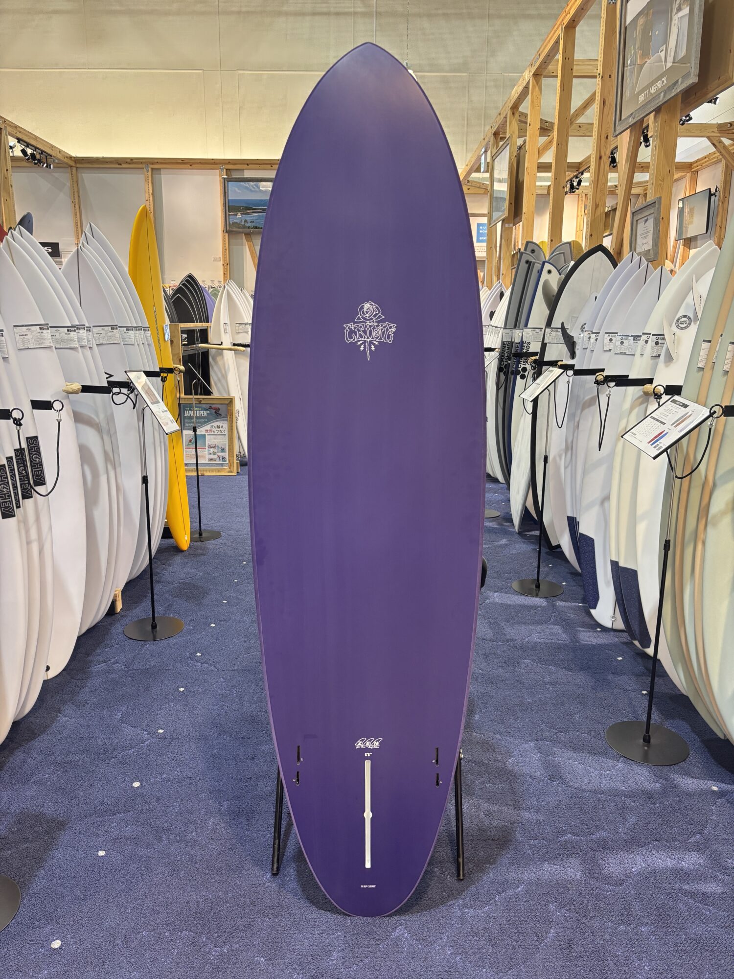 IMG_4178 - SHOP NEWS | ムラサキスポーツオンラインストア 通販 Tall purple surfboard standing upright in a showroom with white and colored boards lined up along wooden racks.