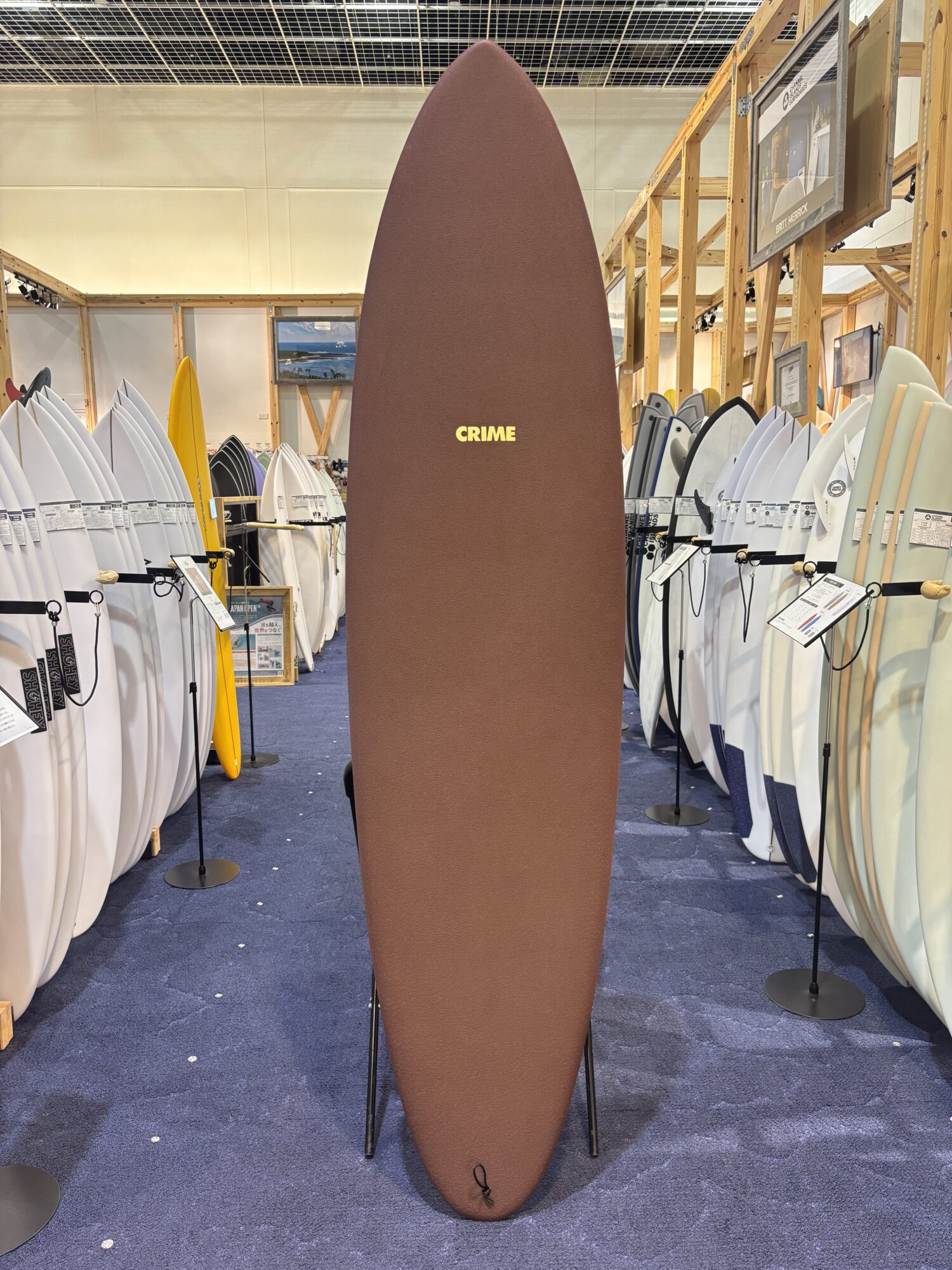 IMG_4179 - SHOP NEWS | ムラサキスポーツオンラインストア 通販 Tall brown surfboard with the word CRIME in yellow letters, standing upright among display boards in a surf shop showroom.