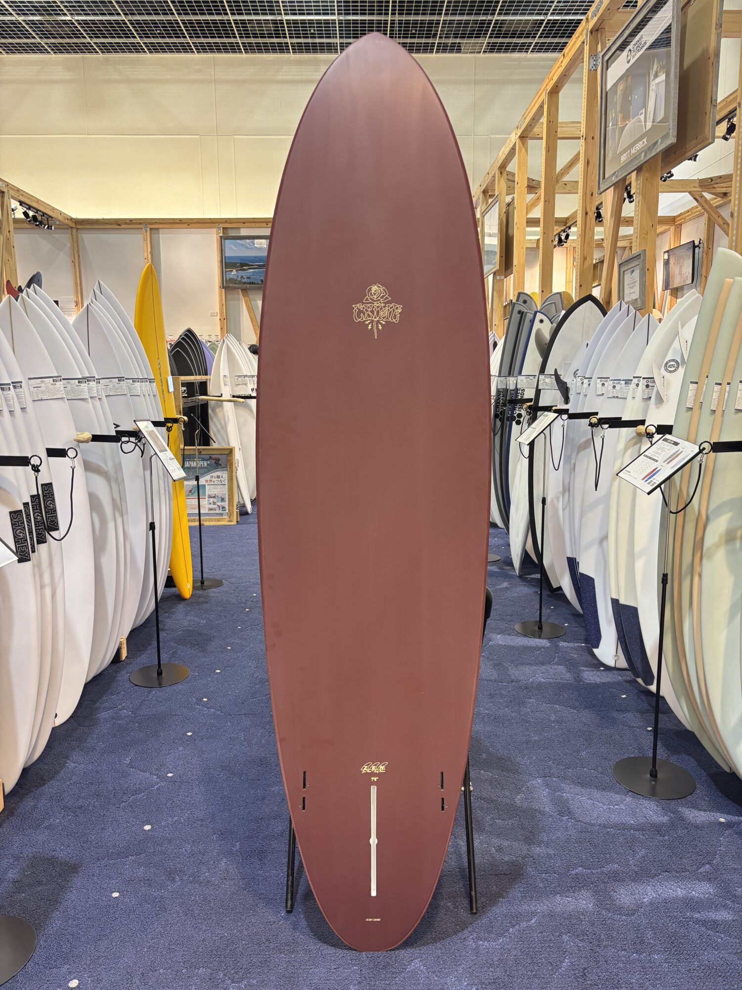 IMG_4180 - SHOP NEWS | ムラサキスポーツオンラインストア 通販 Tall maroon surfboard on display in a shop, surrounded by white and colored boards in a showroom.