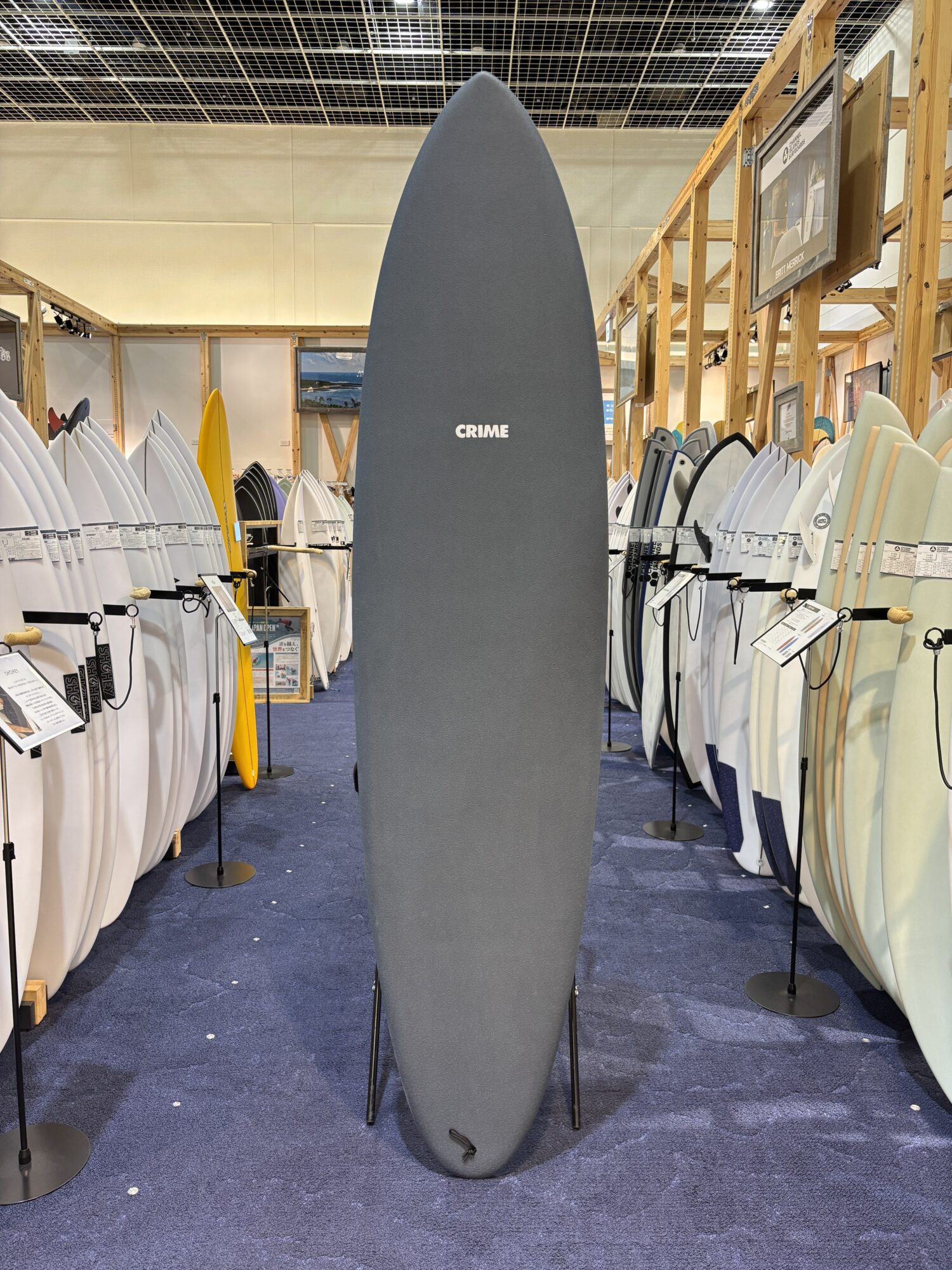 IMG_4181 - SHOP NEWS | ムラサキスポーツオンラインストア 通販 Tall, dark gray surfboard on display in a showroom, surrounded by many white and colored boards on racks.