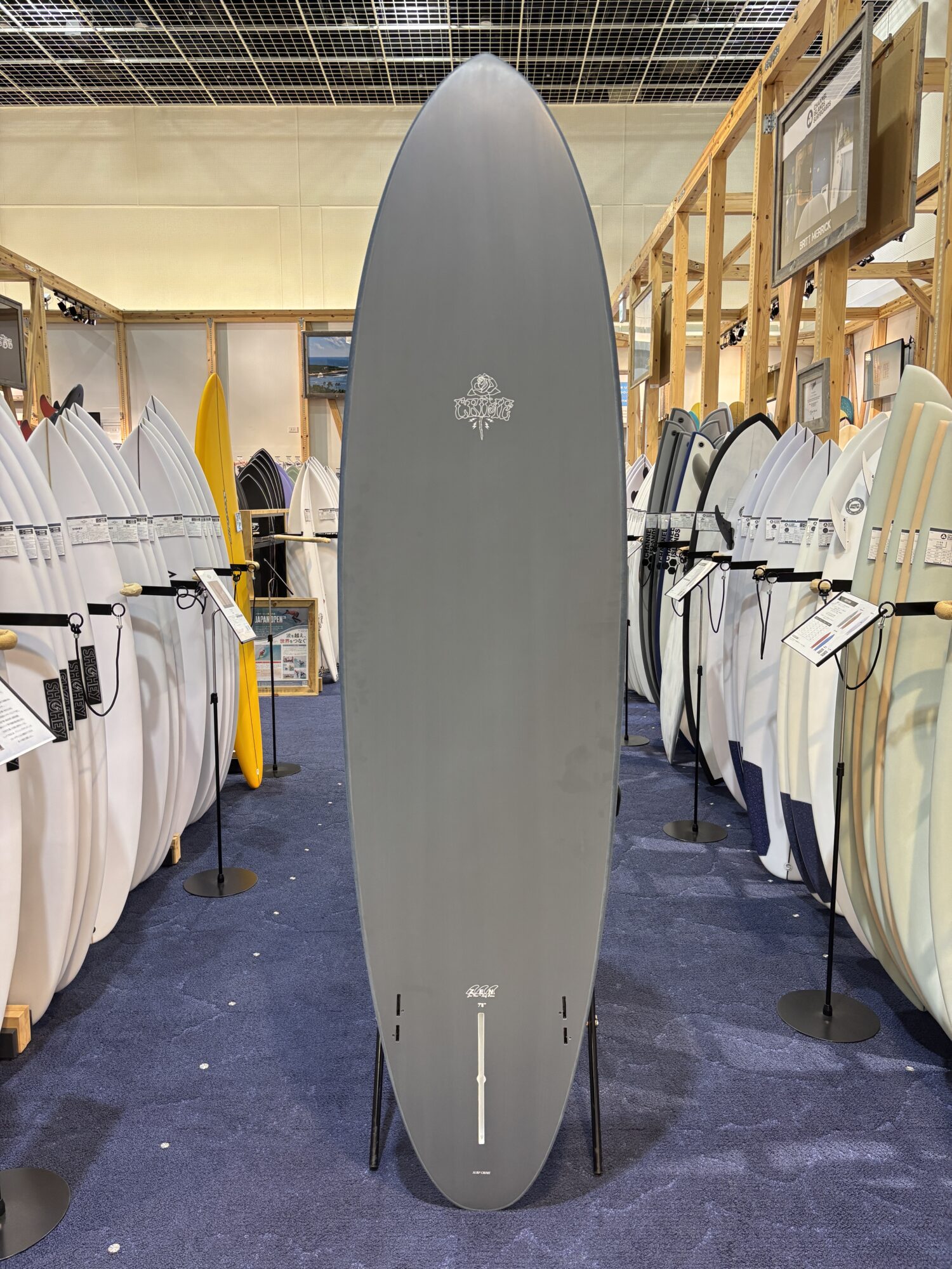 IMG_4182 - SHOP NEWS | ムラサキスポーツオンラインストア 通販 Tall gray surfboard on display in a shop, surrounded by many white and colored boards on racks.