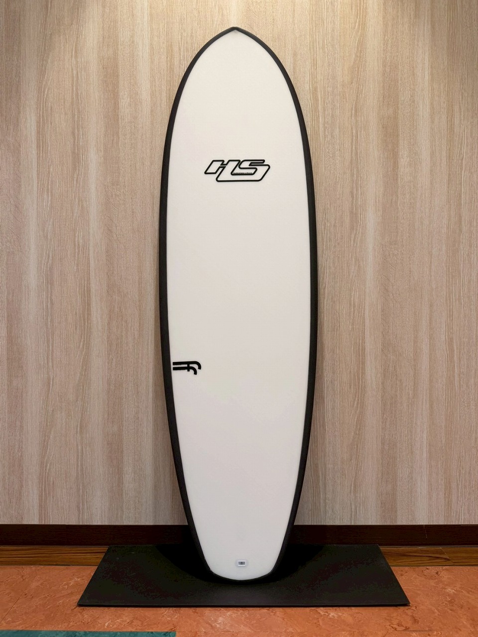 White surfboard with black edging standing upright against a wooden wall on a black display mat.