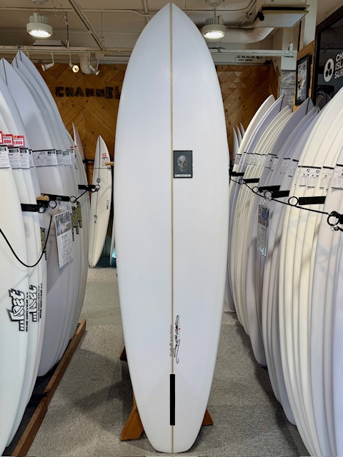 IMG_1864-2 - SHOP NEWS | ムラサキスポーツオンラインストア 通販 White surfboard standing upright on a wooden stand in a surf shop, surrounded by rows of other boards and a small square logo on its chest.