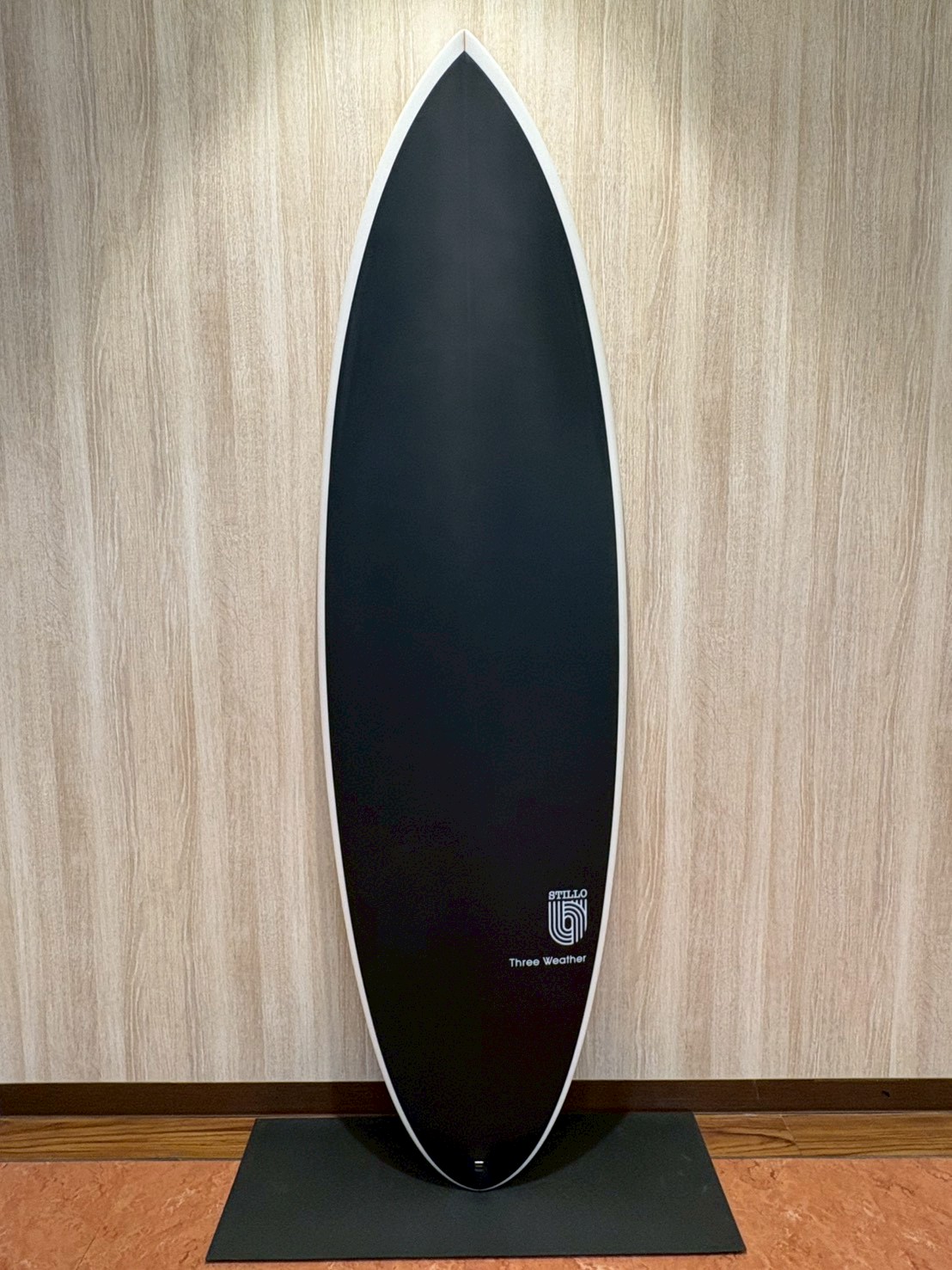LINE_ALBUM_2026 TW_260422_12 - SHOP NEWS | ムラサキスポーツオンラインストア 通販 Vertical black surfboard with white rail, standing upright against a light wood panel wall on a black mat.