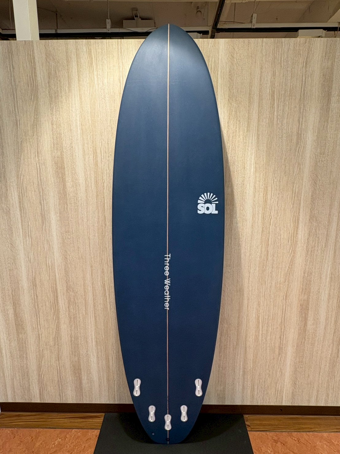 LINE_ALBUM_2026 TW_260422_4 - SHOP NEWS | ムラサキスポーツオンラインストア 通販 Blue surfboard standing upright against a wooden wall, with a centered stringer, five tail fins, and a SOL logo on the right side near the midsection. The vertical text along the stringer reads 'Three Weather'.