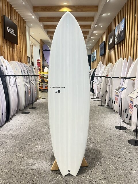 IMG_8570 - SHOP NEWS | ムラサキスポーツオンラインストア 通販 White surfboard standing upright on a display rack in a surf shop, surrounded by rows of other boards on both sides.