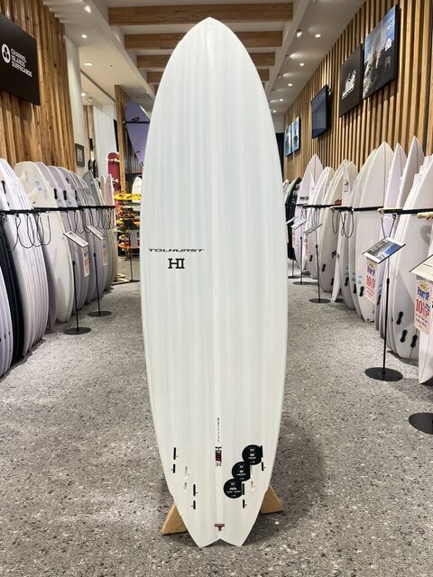 IMG_8571 - SHOP NEWS | ムラサキスポーツオンラインストア 通販 Tall white surfboard on a display stand in a showroom, surrounded by rows of other surfboards against wooden walls.