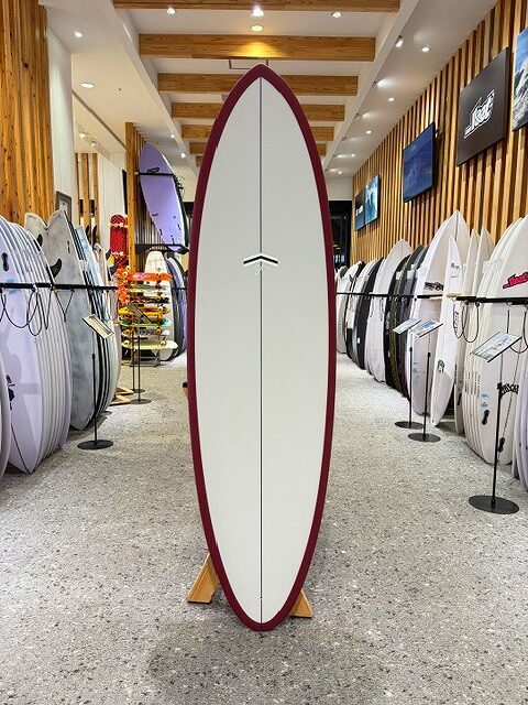 a-IMG_2672 - SHOP NEWS | ムラサキスポーツオンラインストア 通販 White surfboard with a burgundy rim on a display rack in a surf shop, surrounded by other boards on racks in a well-lit showroom.