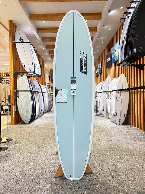 Light blue longboard surfboard on a wooden stand in a surf shop, with other boards on racks along the walls.