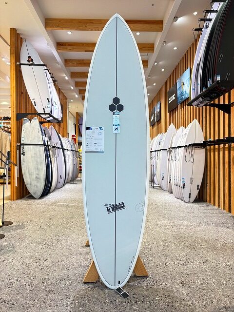 A white surfboard on a stand in a bright showroom, with rows of boards lining the walls behind it.
