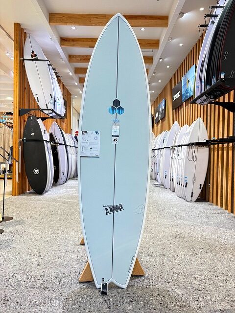 Light blue surfboard on a display stand in a surf shop showroom, with rows of surfboards on racks along wooden walls.