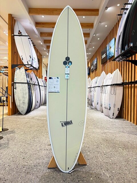 Cream-colored longboard on a display stand in a surf shop, with rows of surfboards lining both walls around it.