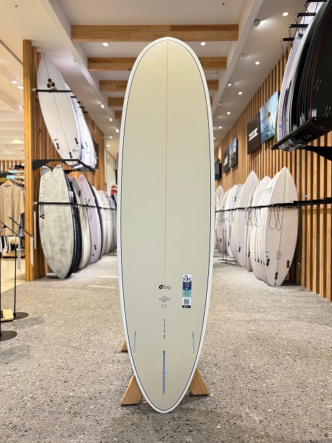 Tall, pale-yellow surfboard on a stand inside a surf shop, surrounded by racks of boards and wooden slat walls.