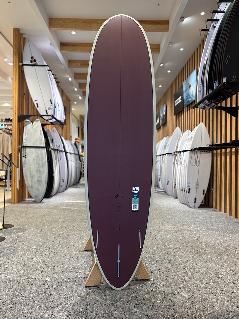 Purple surfboard standing upright on a wooden display stand inside a surf shop, with other boards on racks along the walls.