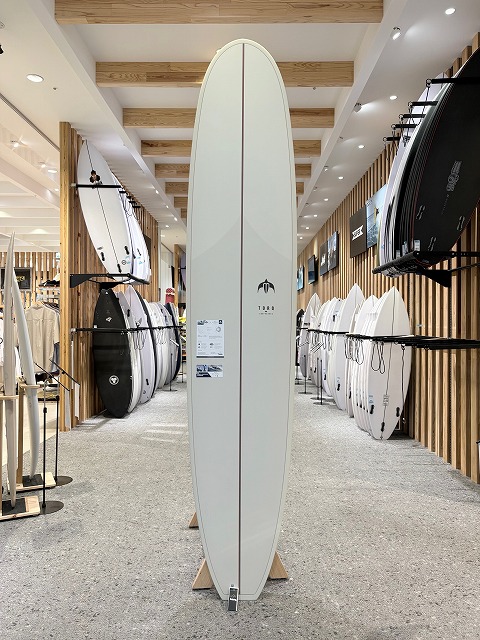 Tall white surfboard on a stand in a surf shop showroom with racks of boards on both sides.