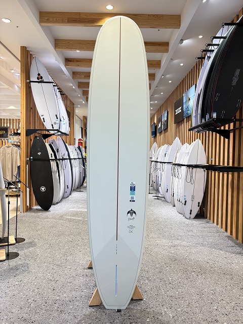 Tall white surfboard with a red stringer standing upright on a wooden rack in a surf shop, surrounded by other boards.