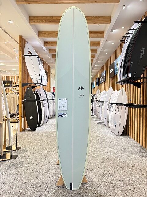 Tall pale green surfboard on a stand in a surf shop showroom, surrounded by other boards on racks.