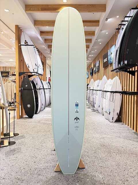Pale mint-green surfboard on a display stand inside a surf shop, with racks of boards along both walls and wood paneling.