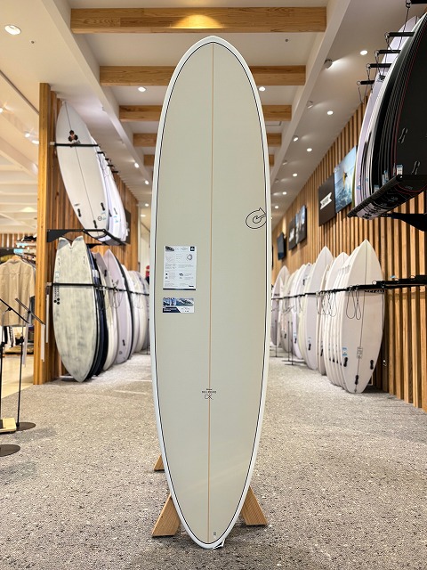 Cream-colored surfboard with a light stringer, standing upright on a display rack inside a bright surf shop.