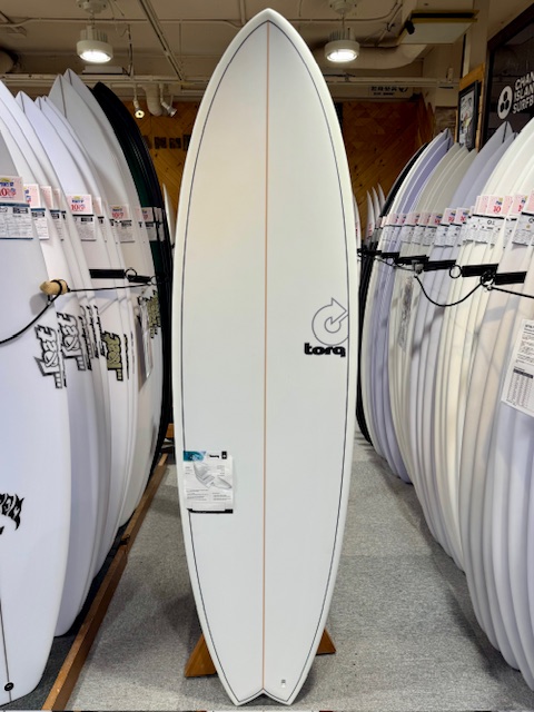 IMG_3689 - SHOP NEWS | ムラサキスポーツオンラインストア 通販 White surfboard displayed upright between rows of other boards in a shop, featuring an orange centerline and a Torq logo on the right side.