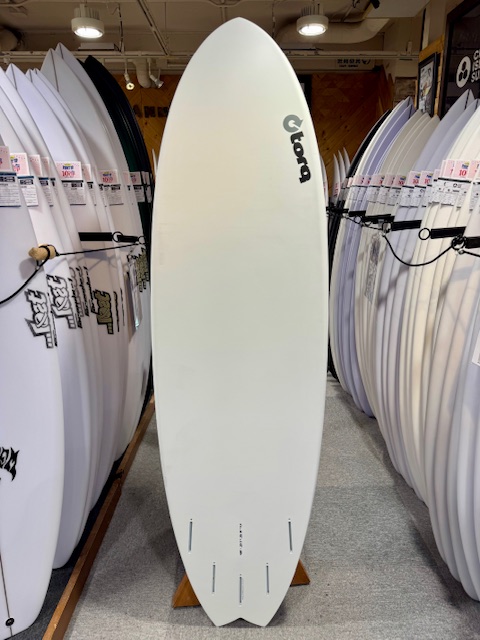 IMG_3691 - SHOP NEWS | ムラサキスポーツオンラインストア 通販 White surfboard on display in a shop, with a Globe logo on the upper right, surrounded by other surfboards on racks.