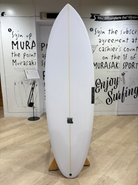 IMG_7099 1 - SHOP NEWS | ムラサキスポーツオンラインストア 通販 White surfboard on a wooden display stand in a showroom, centered against a wall with black handwritten text and drawings in the background.
