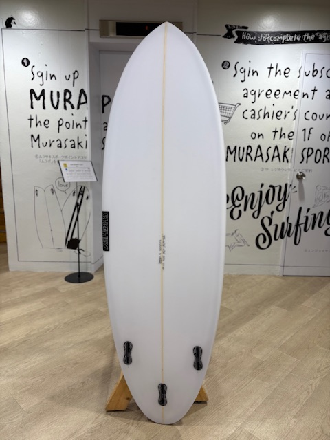 IMG_7100 1 - SHOP NEWS | ムラサキスポーツオンラインストア 通販 White surfboard on a wooden rack indoors, fin boxes visible at the tail, with graffiti-style surf shop walls in the background.