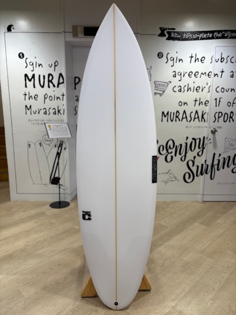 IMG_7103 - SHOP NEWS | ムラサキスポーツオンラインストア 通販 White surfboard standing upright on a wooden rack inside a gallery or showroom, with handwritten wall art in the background.