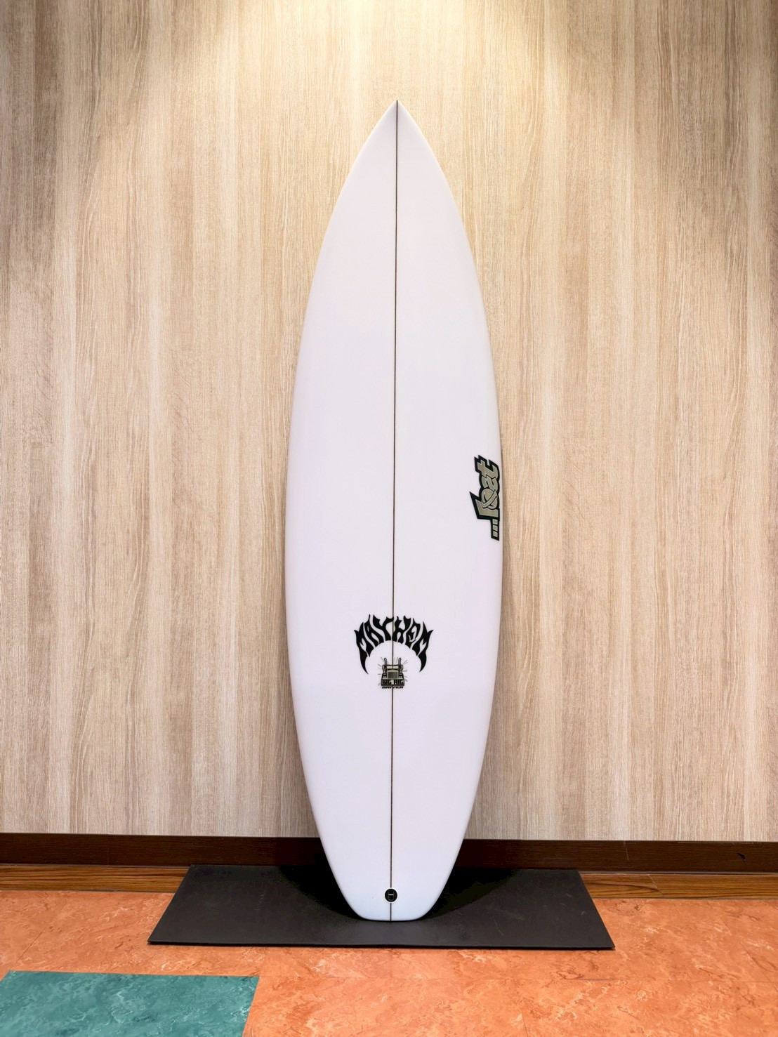 LINE_ALBUM_2026 LOST_260427_1 - SHOP NEWS | ムラサキスポーツオンラインストア 通販 White surfboard standing upright against a wooden wall on a black mat, with black logos near the tail and midboard.