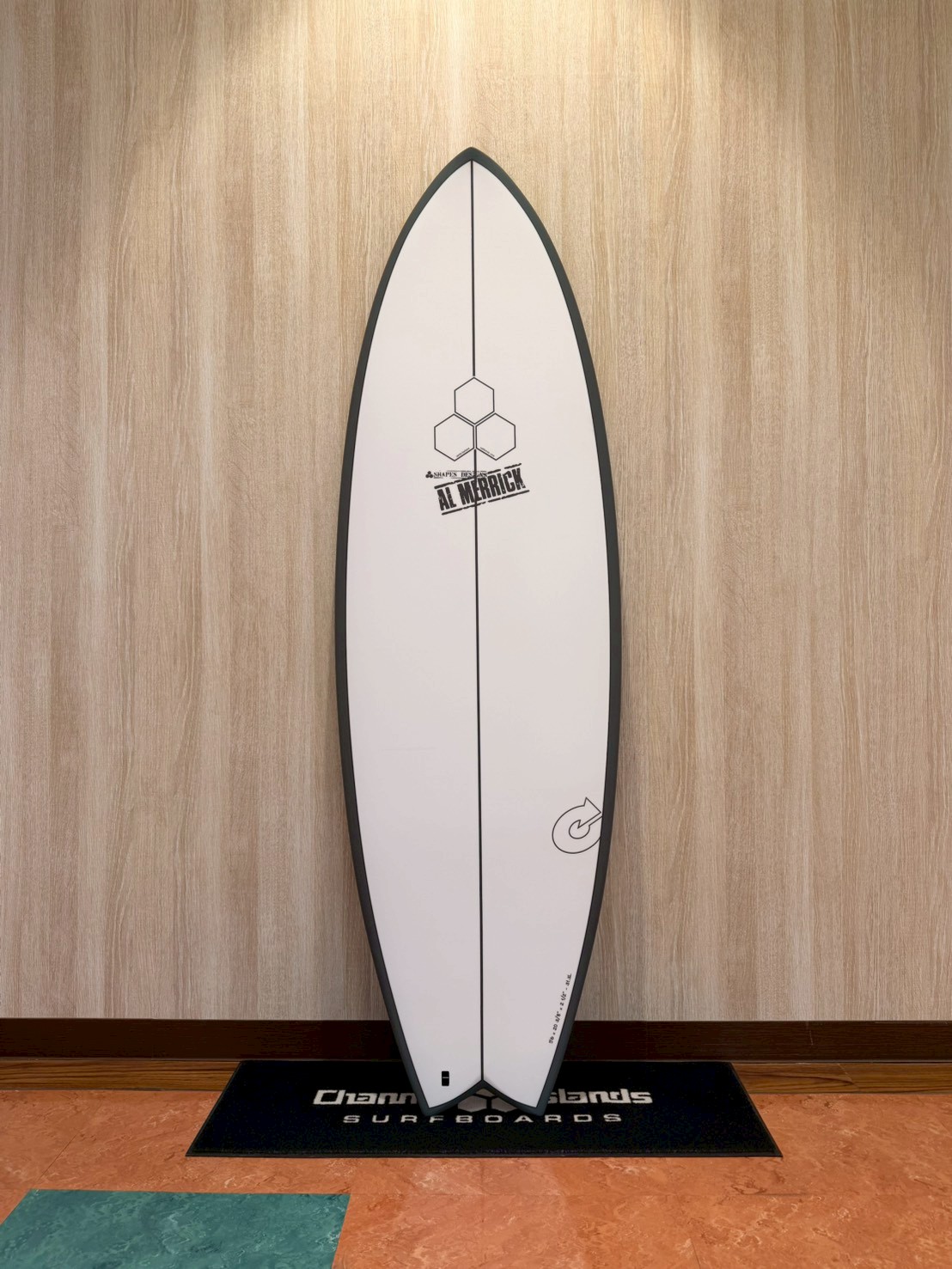 LINE_ALBUM_2026 トルク_260426_3 - SHOP NEWS | ムラサキスポーツオンラインストア 通販 White surfboard with black border standing upright against a wood-paneled wall on a display mat.