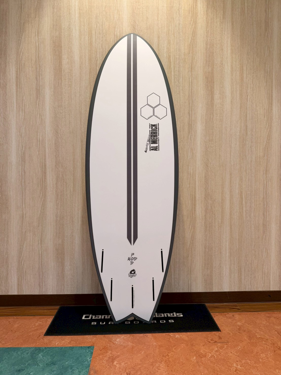 LINE_ALBUM_2026 トルク_260426_4 - SHOP NEWS | ムラサキスポーツオンラインストア 通販 White surfboard with double black center stripes and hexagon motif, leaning against a light wood wall on a black-mat display.