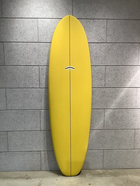 s-IMG_6377 - SHOP NEWS | ムラサキスポーツオンラインストア 通販 Yellow surfboard standing upright against a gray cinder-block wall.