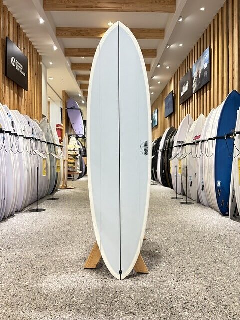 BIG BARON-1 - SHOP NEWS | ムラサキスポーツオンラインストア 通販 White surfboard on a display stand in a surf shop showroom, with rows of other boards lining the walls and bright overhead lighting.