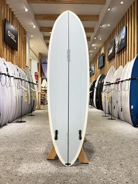 BIG BARON-2 - SHOP NEWS | ムラサキスポーツオンラインストア 通販 White surfboard on a wooden stand in a surf shop, with rows of boards along the walls behind it.