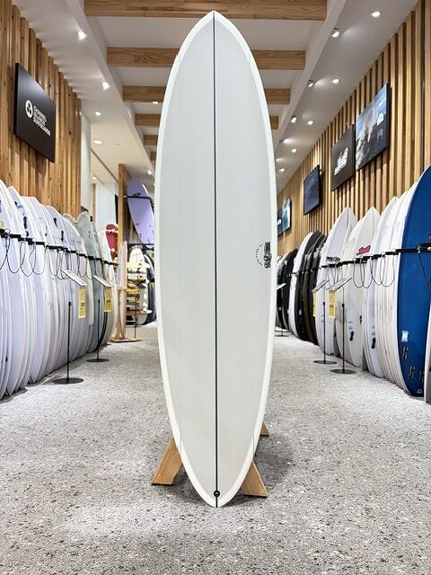 BIG BARON-3 - SHOP NEWS | ムラサキスポーツオンラインストア 通販 Tall white surfboard standing upright on a wooden display in the center of a surf shop, flanked by rows of other boards on racks.