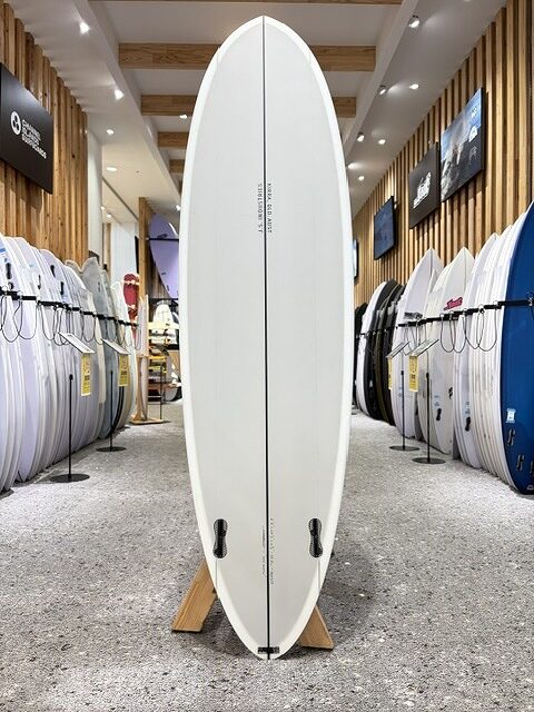 BIG BARON-4 - SHOP NEWS | ムラサキスポーツオンラインストア 通販 White longboard surfboard on a stand in a showroom, with racks of boards along the walls.