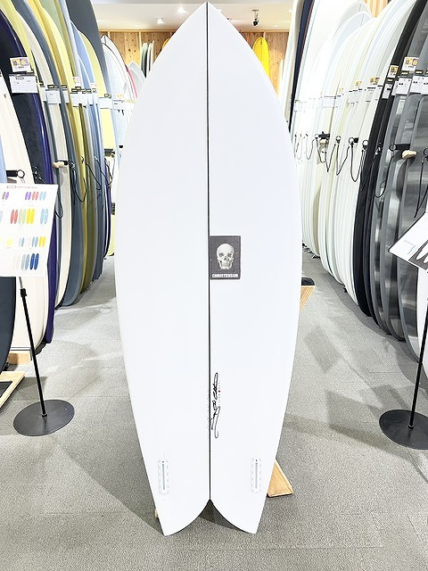 s-IMG_6280 - SHOP NEWS | ムラサキスポーツオンラインストア 通販 White surfboard with a black skull logo stands upright on display among colorful boards in a surf shop showroom.