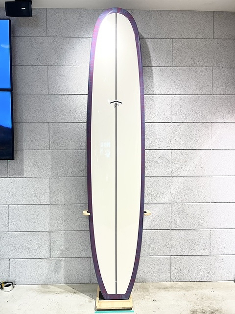 s-IMG_6375 - SHOP NEWS | ムラサキスポーツオンラインストア 通販 Tall surfboard standing upright on a wooden stand against a gray tiled wall; cream center with purple rails.