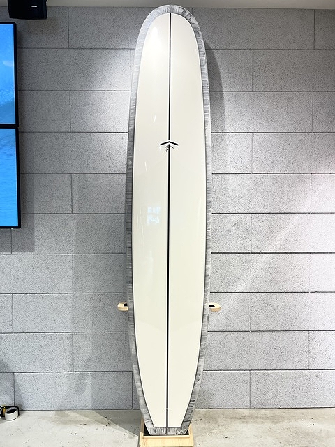 s-IMG_6377 - SHOP NEWS | ムラサキスポーツオンラインストア 通販 Cream-colored surfboard with a black center seam, standing upright on a wooden rack against a gray brick wall.