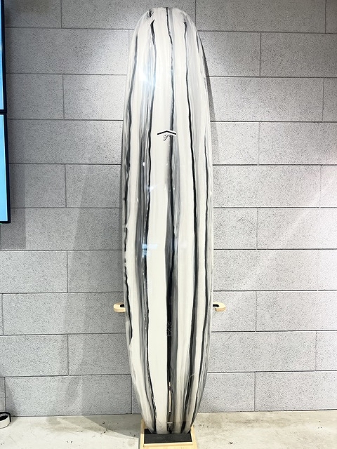 s-IMG_6378 - SHOP NEWS | ムラサキスポーツオンラインストア 通販 Tall white surfboard with black vertical stripes, standing upright on a wooden rack against a gray wall in a showroom setting.