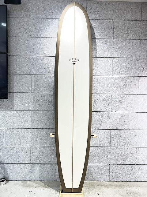 A white surfboard with a dark brown edge standing upright against a gray tiled wall on a wooden stand, no fins visible.