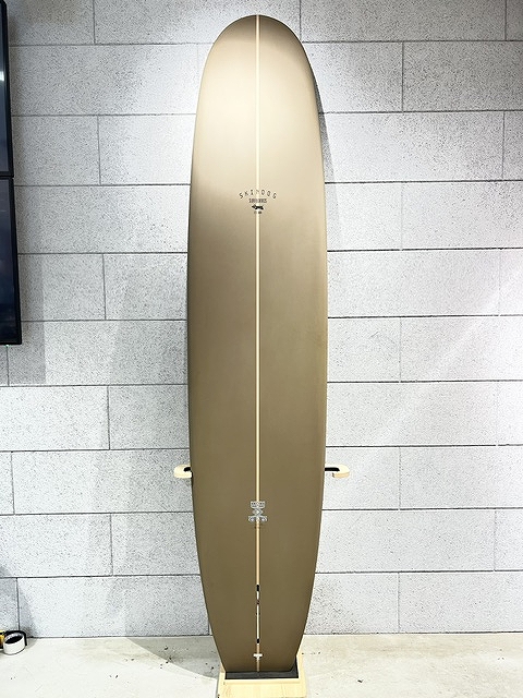 Brown longboard surfboard standing upright on a wooden display stand against a gray brick wall, center stringer visible down the board.